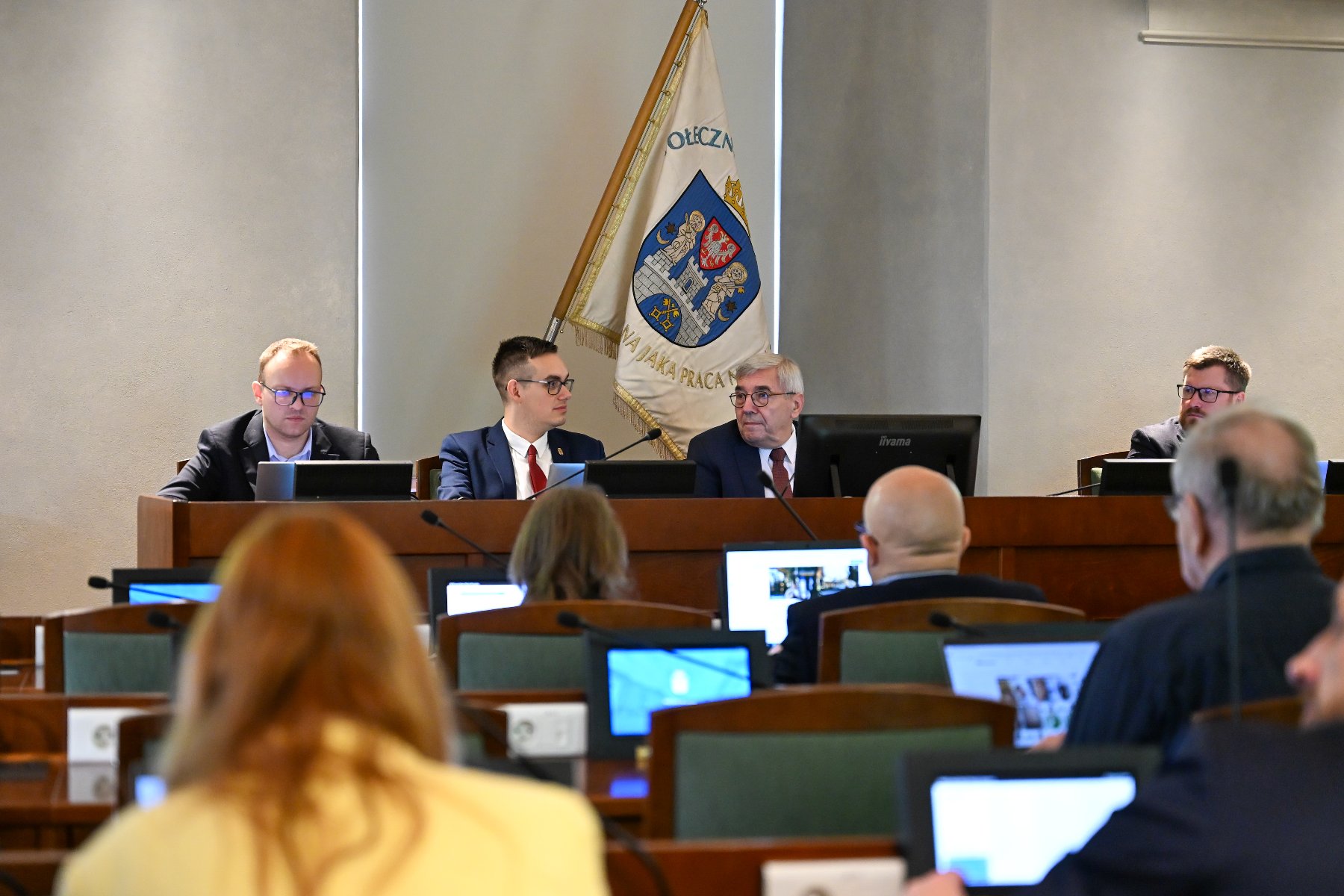Widok na salę sesyjną podczas posiedzenia. Na podwyższeniu siedzi czterech mężczyzn w garniturach, patrzących przed siebie. W tle wisi biała flaga z herbem i napisem, częściowo widocznym. Na pierwszym planie, rozmazana, widoczna jest publiczność, której część patrzy w ekrany laptopów i monitorów.