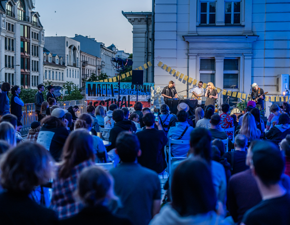 Podczas festiwalu sztuka wyjdzie na ulice Poznania. Widownia stoi zwrócone do sceny, na której występuje zespół muzyczny. Występ odbywa się przed białym, zabytkowym budynkiem. Obok sceny powieszono duży baner z nazwą wydarzenia. W tle rozciągają się kolejne budynki kamienic.