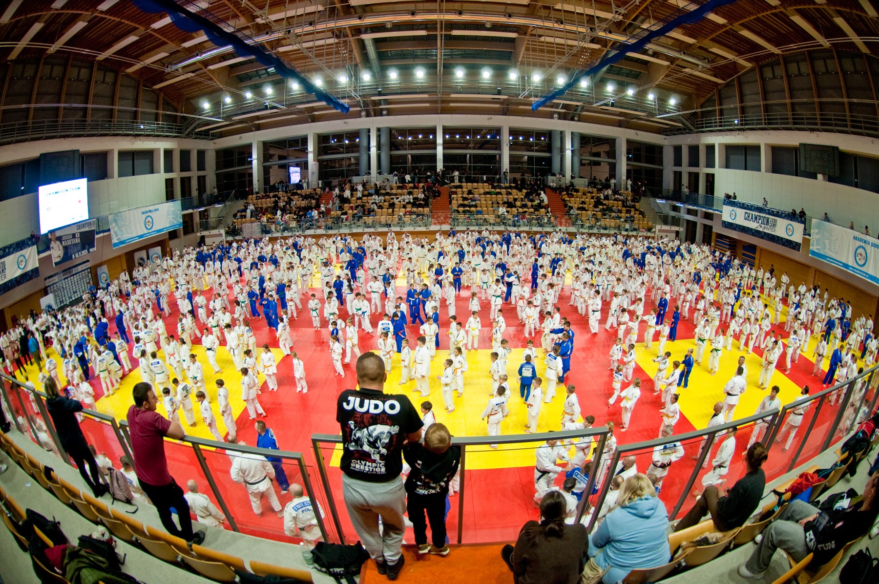 Winter Champion Judo Camp, fot. Facebook Champion Judo Camp - na macie widać dużo ludzi w niebieskich i białych strojach, którzy biorą udział we wspólnym treningu. Oprócz nich widać ludzi na trybunach, którzy obserwują zmagania na macie