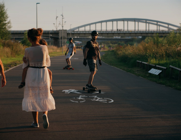 Zachód słońca nad Wartą. Trasa wartostrady. Na pierwszym planie dwie kobiety. Jedna z nich trzyma dziecko. Na drugim planie dwóch mężczyzn jedzie na deskorolce. W tle most św. Rocha.