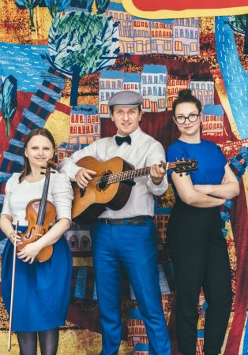 Zdjęcie trzyosobowego zespołu muzycznego (gitara, skrzypce i wokal). W tle namalowany kolorowy plan miasta.