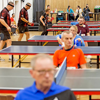 zdjęcie z turnieju tenisa stołowego; na zdjęciu widoczni grający zawodnicy i przypatrujący się grze sędziowie
