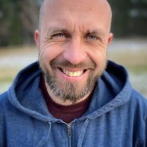 Zdjęcie Roberta Maciąga. Stoi uśmiechnięty. Głowa łysa, niebieska bluza z opuszczonym na ramiona kapturem.