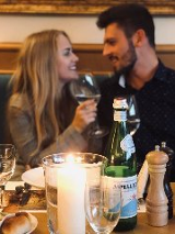 Usmiechnięta para siedzi na kanapie przy stole, na którym stoją kieliszki, szampan i róże w wazonie.