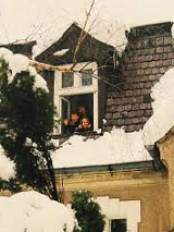 Na zdjęciu widać ośnieżony dach domu z małym wysuniętym oknem mansardowym. W otwartym oknie stoją trzy uśmiechnięte dzieci, które wyglądają na zadowolone i machają w stronę fotografa. Dach pokryty jest grubą warstwą śniegu, a gałęzie drzew wokół również są przysypane. Scena sprawia wrażenie zimowego, rodzinnego i radosnego momentu.