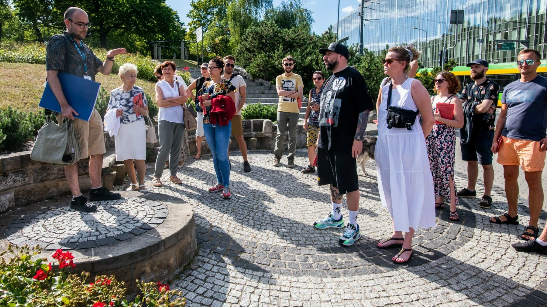 Ludzie stojący na placyku przy parku Dąbrowskiego przy Górnej Wildzie słuchający przewodnika w słoneczny dzień. Za nimi tramwaj i nowoczense budownictwo.