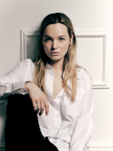 Zdjęcie Michaliny łabacz - młodej kobiety, w białej bluzce i czarnych spodniach, siedzacej pod ścianą, z jedną nogą zgiętą w kolanie.