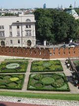 Francuskie ogrody otoczone ceglastym murem, za nim kamienice i ulica. Widok z góry.