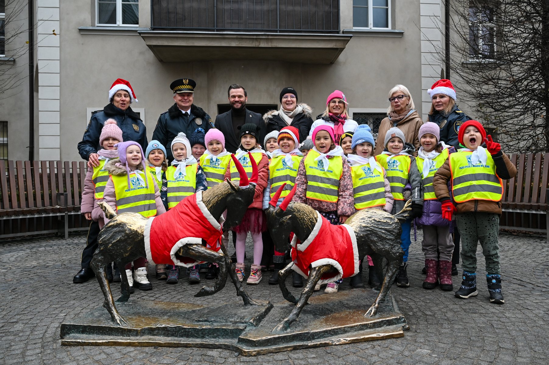 Zdjęcie przedstawia dzieci ubierające koziołki i stojących obok nich dorosłych, w tym dziennikarzy z mikrofonami i kamerami.
