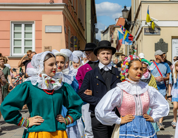 Zdjęcie przedstawia grupę ludzi na Starym Rynku przebranych w tradycyjne bamberskie stroje, m.in. kolorowe sukienki i ozodby z kwiatów lub białe czepki na głowie. Za nimi widać tłum innych ludzi.