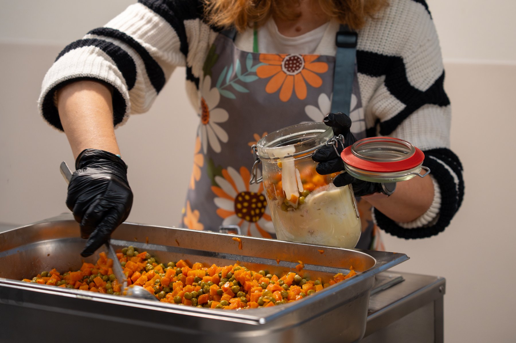 Zdjęcie przedstawia kobietę nakładającą jedzenie do słoika. W misce przed nią znajdują się pokrojony groszek i marchewka.