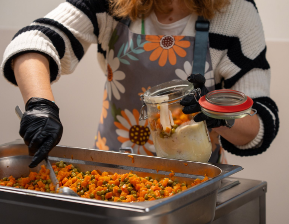 Zdjęcie przedstawia kobietę nakładającą jedzenie do słoika. W misce przed nią znajdują się pokrojony groszek i marchewka.