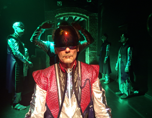 Zdjęcie przedstawia mężczyznę w średniowiecznym stroju z dziwnym kaskiem na głowie. W tle widać innych mężczyzn.