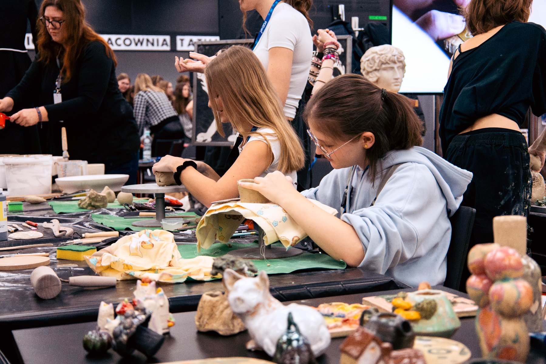 Zdjęcie przedstawia młodą dziewczynę rzeźbiącą w kamieniu stojącym na wysokim stołku.