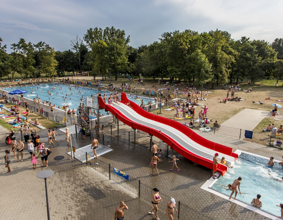 Kąpieliska w sezonie 2024 Zdjęcie przedstawia odkryty basen. Mnóstwo ludzi pływających, dzieci zjeżdżają na dużej zjeżdżalni. Wokół są drzewa.