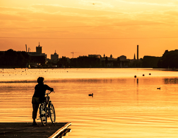 Zdjęcie przedstawia osobę stojącą na rowerze na pomoście przy jeziorze Maltańskim, patrzącą w kierunku zachodzącego słońca.