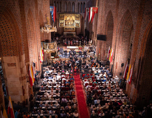 Zdjęcie przedstawia wnętrze katedry. Widać na nim ludzi siedzących w ławkach, a w miejscu ołtarza muzyków z instrumentami. Zdjęcie zrobiono z góry.