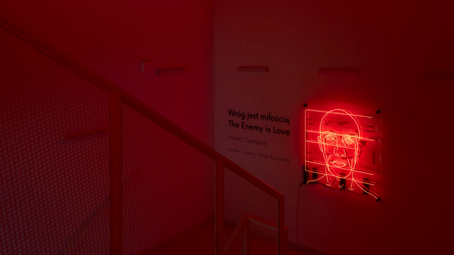 Zdjęcie schodów oraz ścian w Galerii. Na ścianie wisi neon przedstawiający portret mężczyzny ułożony z neonowych linii i kształtów, podświetlony na czerwono-pomarańczowo. Obok widać tytuł wystawy i nazwisko autora.