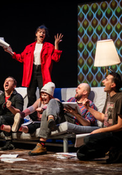 na kanapie na scenie siedzi kilka osób, niektórzy trzymają w rękach otwarte książki. Za nimi stoi kobieta z otwartą książką w ręce, mocno gestykuluje