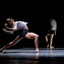 Tancerka w białej koszuli i czarnych szortach w tanecznej pozie - wygięta do tyłu, z jedną nogą zgięta, a drugą wyprostowaną. W tle druga osoba stoi z ugiętą nogą, czarne tło.