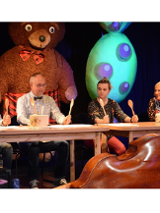 Na scenie czworo aktorów siedzi za stołem. W rękach trzymają uniesione drewniane łyżki, a na stołach leżą odwrócone do góry dnem garnki.