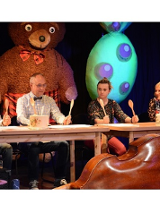 Na scenie czworo aktorów siedzi za stołem. W rękach trzymają uniesione drewniane łyżki, a na stołach leżą odwrócone do góry dnem garnki.