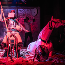 Mężczyzna z zakrytą twarzą siedzi na drewnianym siedzisku przypominającym siodło, trzyma w ręce długi kij. Po prawej stronie figura konia do połowy zakopana w piasku.