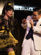 na zdjęciu mężczyzna ubrany w biały garnitur stoi unosząc ręce przed siebie przed krzyczącą na niego kobietą, ubraną w żółto-czarną sukienkę