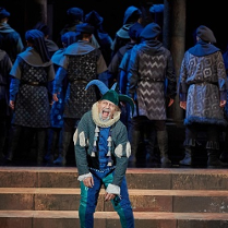 Błazen w zielono-niebieskim stroju i błazeńskiej czapce na scenie, za nim odwróceni tyłem do niego aktorzy, wszyscy w podobnych strojach i nakryciach głowy.