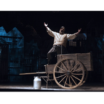 Mężczyzna ubrany w jasną szeroką koszulę, kamizelkę i czarne spodnie stoi na drewnianym wozie. Śpiewa wyciągając ręce przed siebie. Przed wozem stoi kanka na mleko.