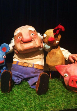 Lalka przedstawiająca starszego mężczyzny obejmującego myszkę, koguta i świnkę. Czarne tło.