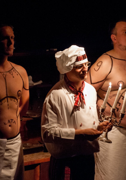 Na zdjęciu trzech mężczyzn: jeden, w środku, ubrany w biały uniform z biała kucharską czapką na głowie, trzyma w ręce świecznik, dwóch pozostałych z nagimi wytatułowanymi torsami za nim.