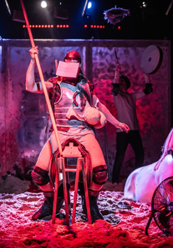 Mężczyzna z zakrytą twarzą siedzi na drewnianym siedzisku przypominającym siodło, trzyma w ręce długi kij. Po prawej stronie figura konia do połowy zakopana w piasku.