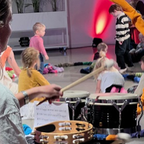 Na pierwszym planie kobieta grająca na perkusji odwrócona tyłem do obiektywu, na drugim planie kilkoro dzieci bawiących się na podłodze.