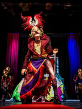 Na zdjęciu postać ubrana w kolorowy strój z maską na twarzy. Stoi na scenie na pierwszym planie, za nią w tle inne postacie.