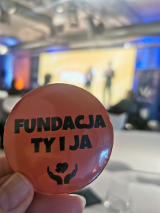 Dłoń trzymająca przypinkę z napisem "Fundacja ty i ja". Tło rozmazane.