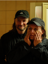 Dwóch mężczyzn w ciemnych ubraniach i czapkach z daszkiem stoi w słabo oświetlonym pomieszczeniu. Mężczyzna z przodu trzyma dłonie na policzkach, wyglądając na zmęczonego lub zrezygnowanego, podczas gdy drugi, stojący za nim się uśmiecha..