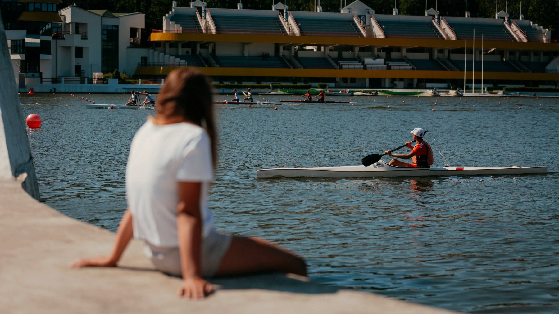 Zdjęcie przedstawia osobę patrzącą na Jezioro Maltańskie, na którym widać kajakarzy, a w tle trybuny zespółu sportowo-rekreacyjnego.