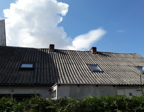 Obraz przedstawia dom z dachem z blachy falistej, na którym znajdują się dwa świetliki i dwa kominy. Dom jest częściowo zasłonięty przez żywopłot na pierwszym planie. Na niebie widać kilka chmur, ale jest ono w większości bezchmurne, co sugeruje przyjemną pogodę. - grafika artykułu