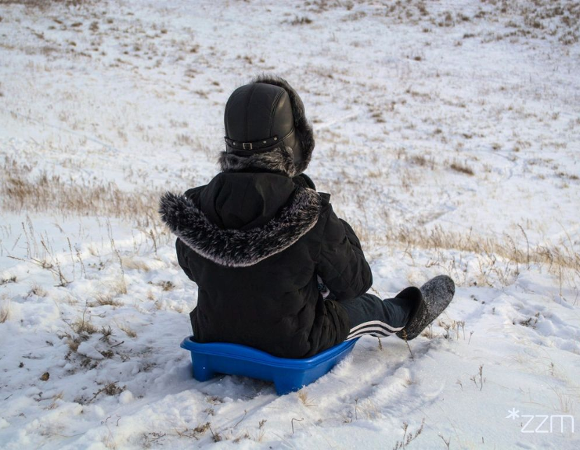 Dziecko ubrane w czarną, puchową kurtkę siedzi na niebieskim siedzisku i zjeżdża z górki. Na ziemi leży biały śnieg.