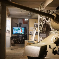 Ekspozycja w muzeum: po lewej na scianie monitor z wyswietlona postacią, w dłebi duży monitor, po prawej czołg i duże zdjęcia na scianie postaci.
