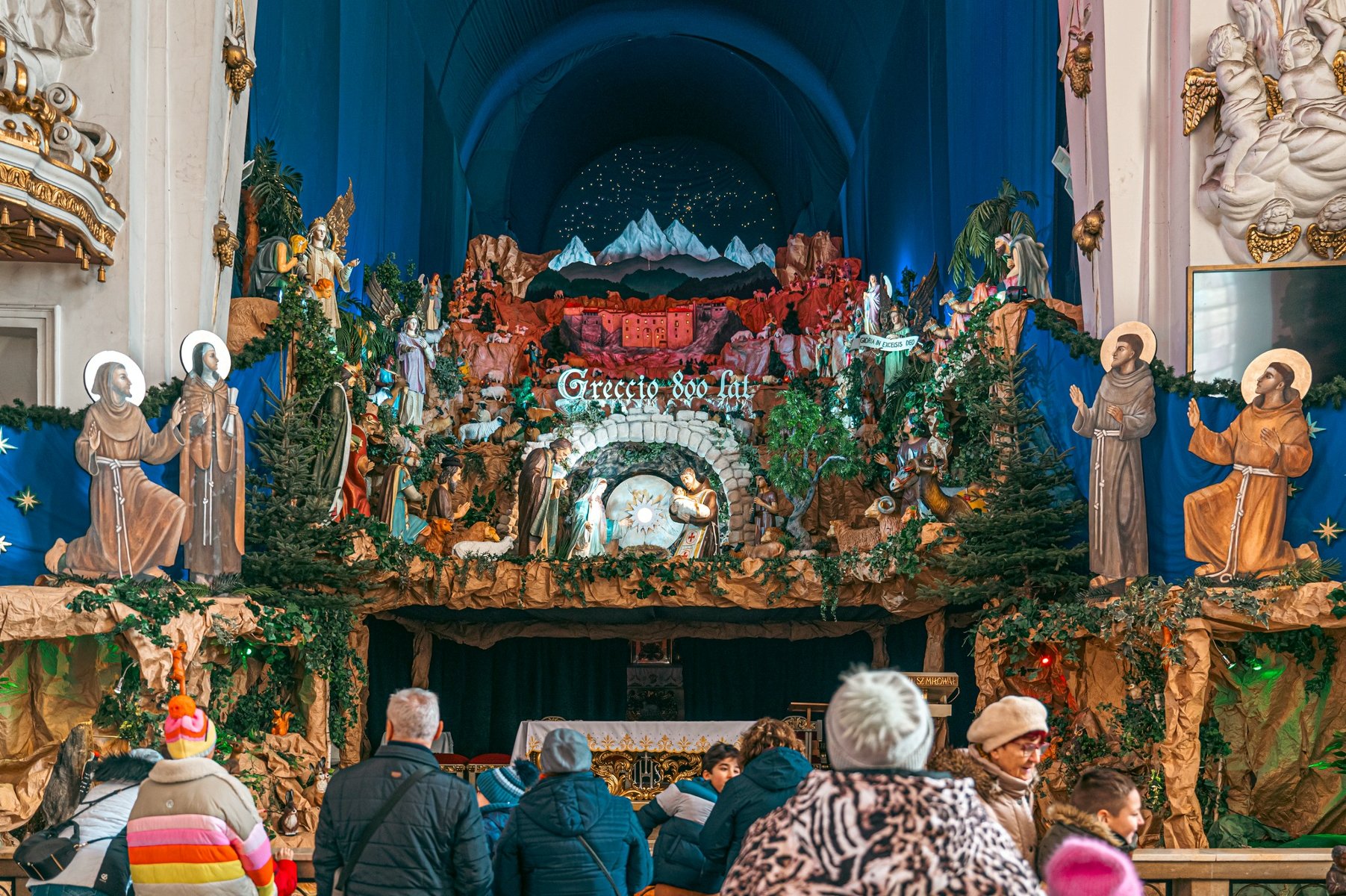 Szopka bożonarodzeniowa na miejscu ołtarza w kościele. Przed ołtarzem stoją ludzie w kurtkach i czapkach. - grafika artykułu