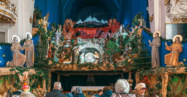 Szopka bożonarodzeniowa na miejscu ołtarza w kościele. Przed ołtarzem stoją ludzie w kurtkach i czapkach.