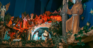 Zdjęcie szopki bożonarodzeniowej z dołu. Figury Józefa, Marii i Jezusa, w tle duże figury apostołów. Obok Trzej Królowie i inne postacie biblijne.