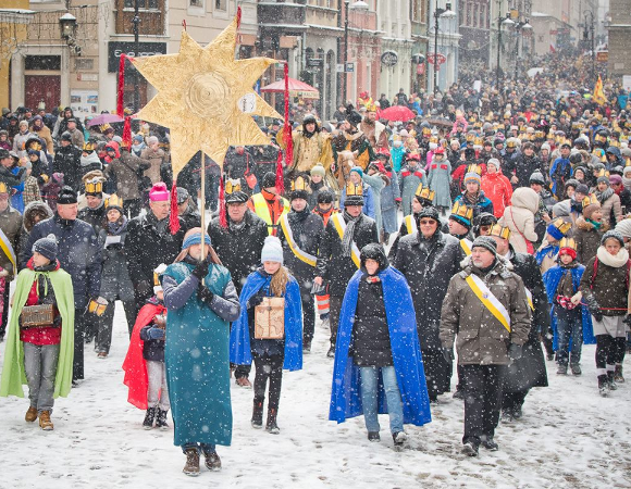 Pochód ludzi ubranych w zimowe stroje i kolorowe peleryny. Na czele idzie osoba trzymająca sztandar ze słońcem. Pada śnieg, na chodniku leży śnieg. W tle kamienice.