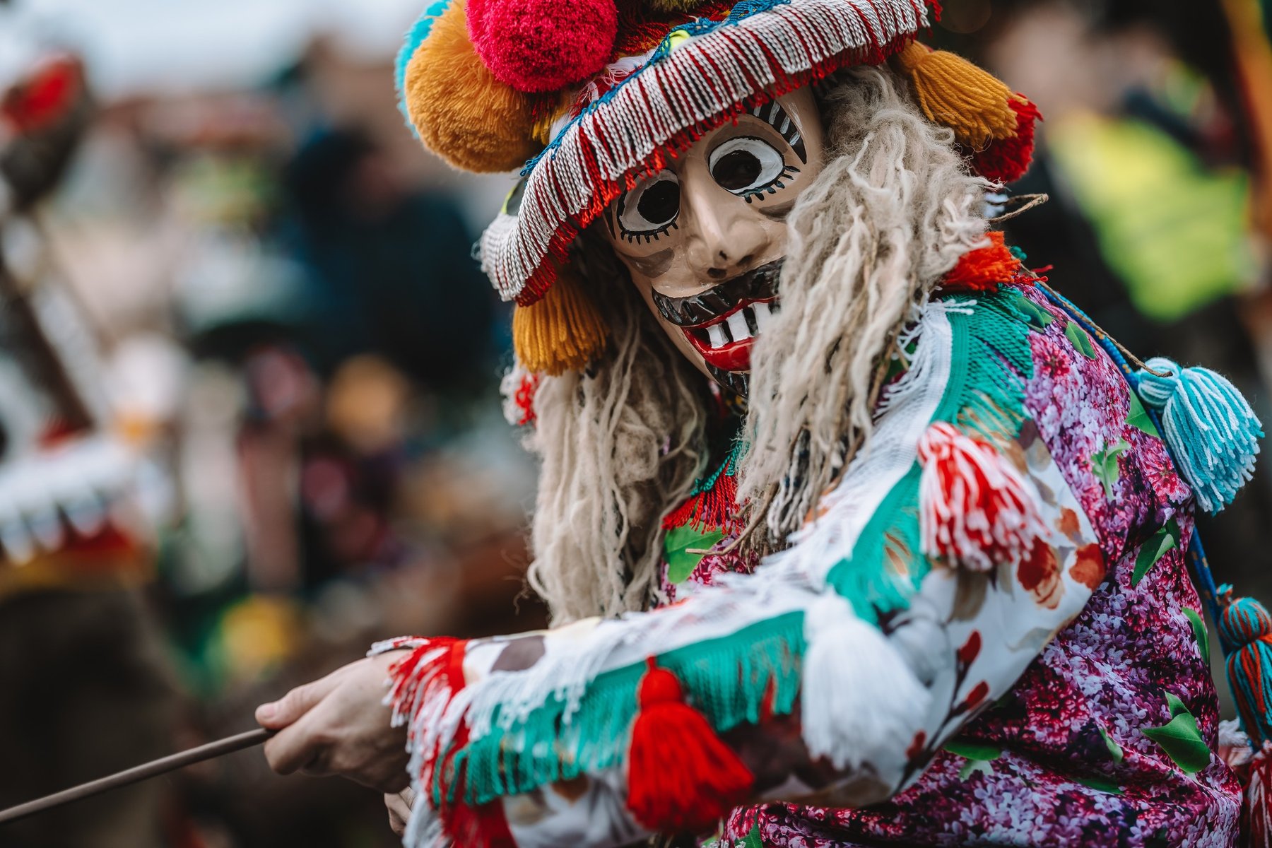 W sobotę korowód Dziadów przejdzie ulicami Starego Miasta / fot. Regionalny Ośrodek Kultury w Bielsku-Białej Postać w tradycyjnym stroju ludowym nosi maskę o wyrazistych, malowanych rysach twarzy z dużymi oczami i szerokim uśmiechem ukazującym zęby. Maska jest otoczona długimi, jasnymi włosami z wełny. Na głowie ma ozdobny kapelusz z frędzlami, pomponami i kolorowymi dodatkami. Strój z kwiecistej koszuli, haftowanych elementów i frędzli w czerwonym, zielonym oraz białym kolorze. W rozmytym tle uczestnicy wydarzenia.